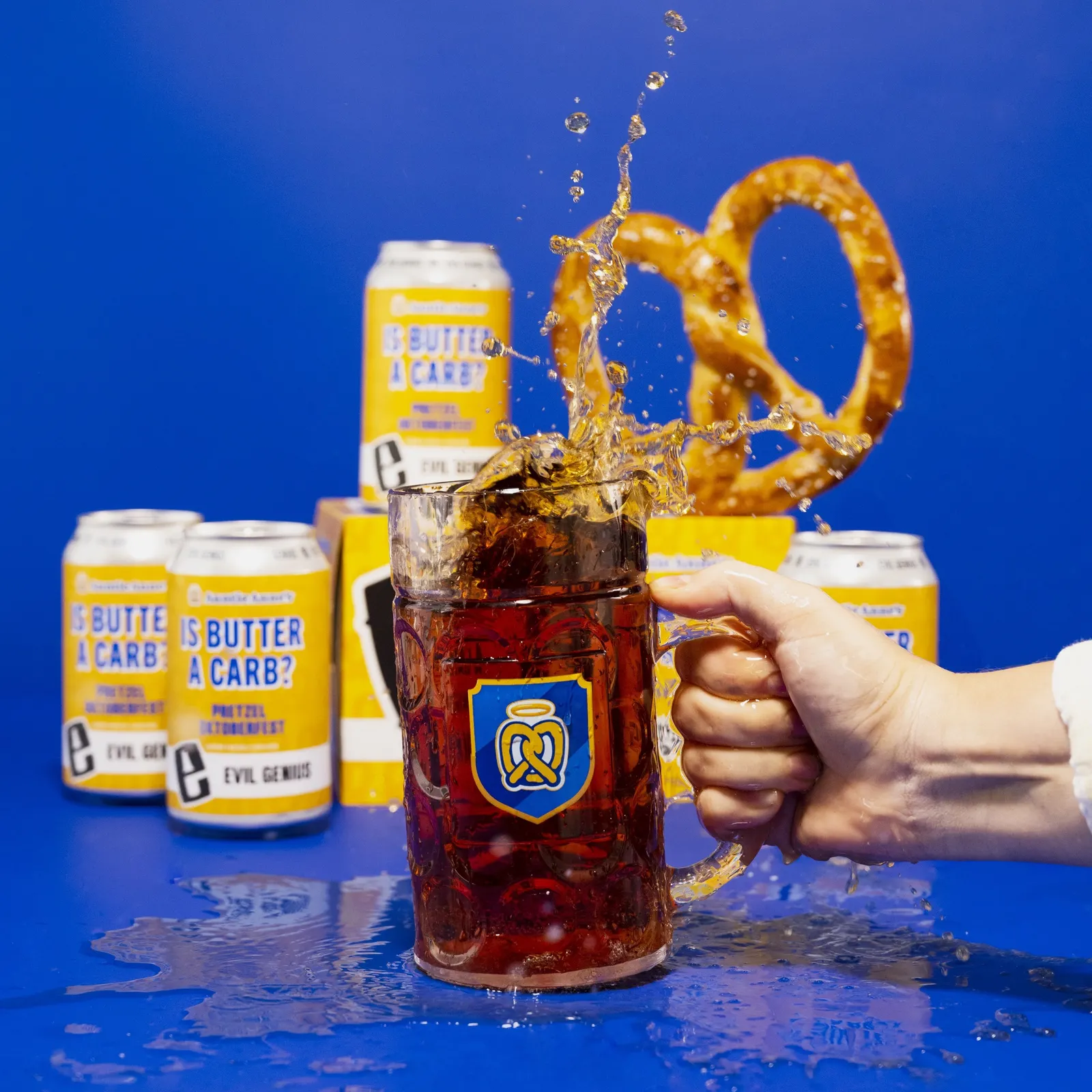 A hand holds a full glass beer stein that is sloshing over the edge, with yellow "Is Butter a Carb?" cans and an Auntie Anne's soft pretzel in the background.