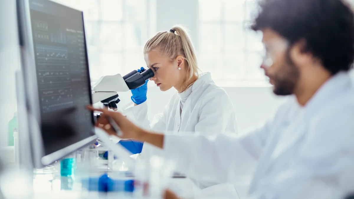 A male researcher analyzes data on a computer as a female colleague analyzes a specimen under a microscope.