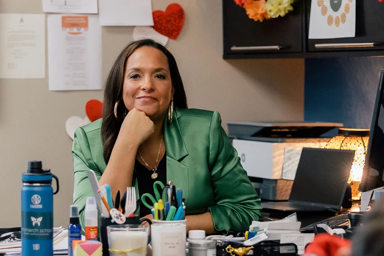 A person is sitting behind a desk, looking at the camera. On the desk are several items including a water bottle and computer monitors. Another person can be seen in the forefront out of focus.