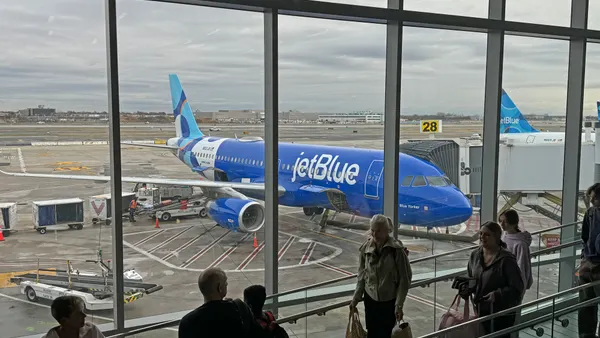 Travelers line up in and airport, a window behind them shows a JetBlue plane.