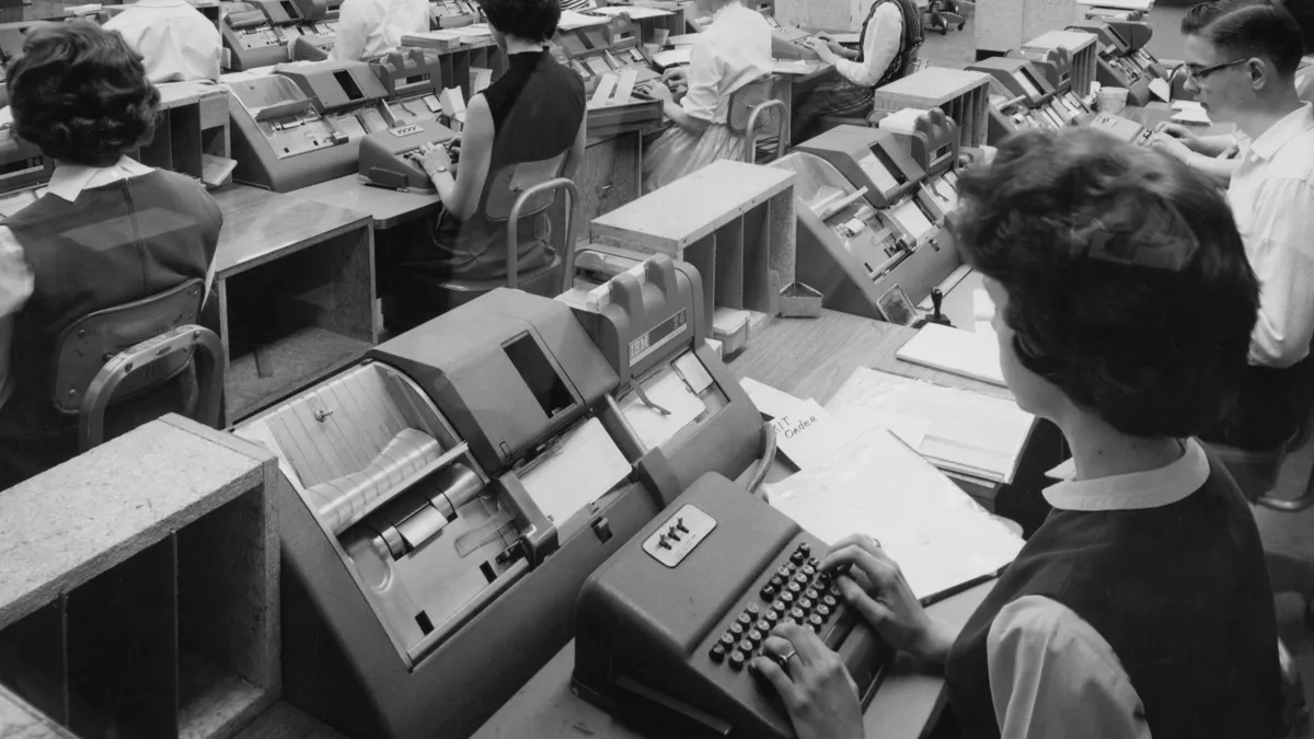 A full office of card punch operators are hard at work in this black and white image, from circa 1965