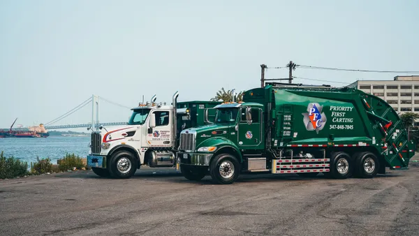 Two trash trucks with Priority First Carting branding sit on a concrete lot near the water, with the Verrazzano-Narrows Bridge to Staten Island visible behind them.