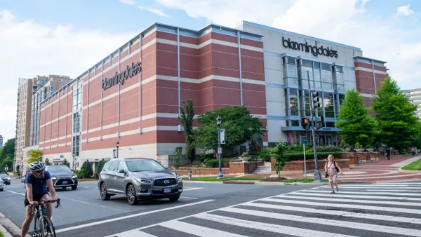 Traffic including cars and a cyclist ride by a Bloomingdale's store under blue skies, as a nearby pedestrian crosses the street.
