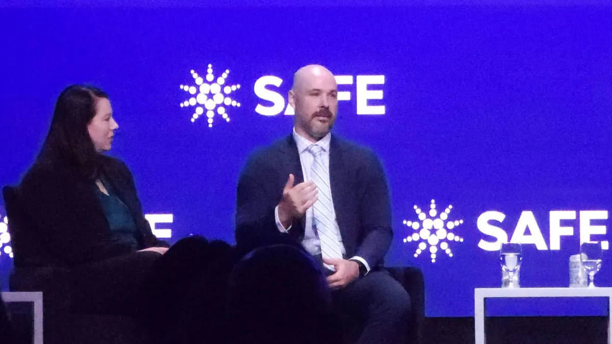 A woman in a lback suit looking at a man in a gray suit talking to the audience on stage.