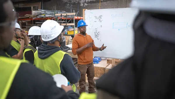 A man in a blue construction hat teaches apprentices at a workshop, gesturing in front of a white board.