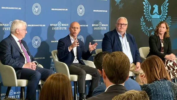 Four people are sitting in chairs on a stage. One person it talking. There is a blue background with wording.