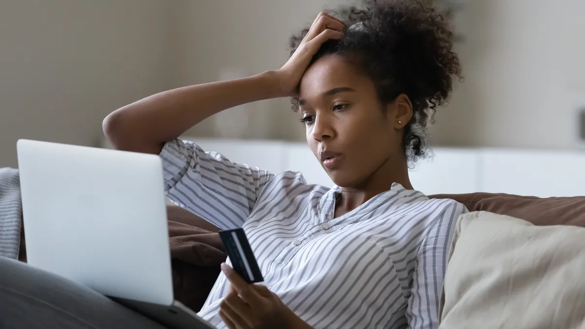 A young woman holding a credit card with a laptop on her legs looking stressed.