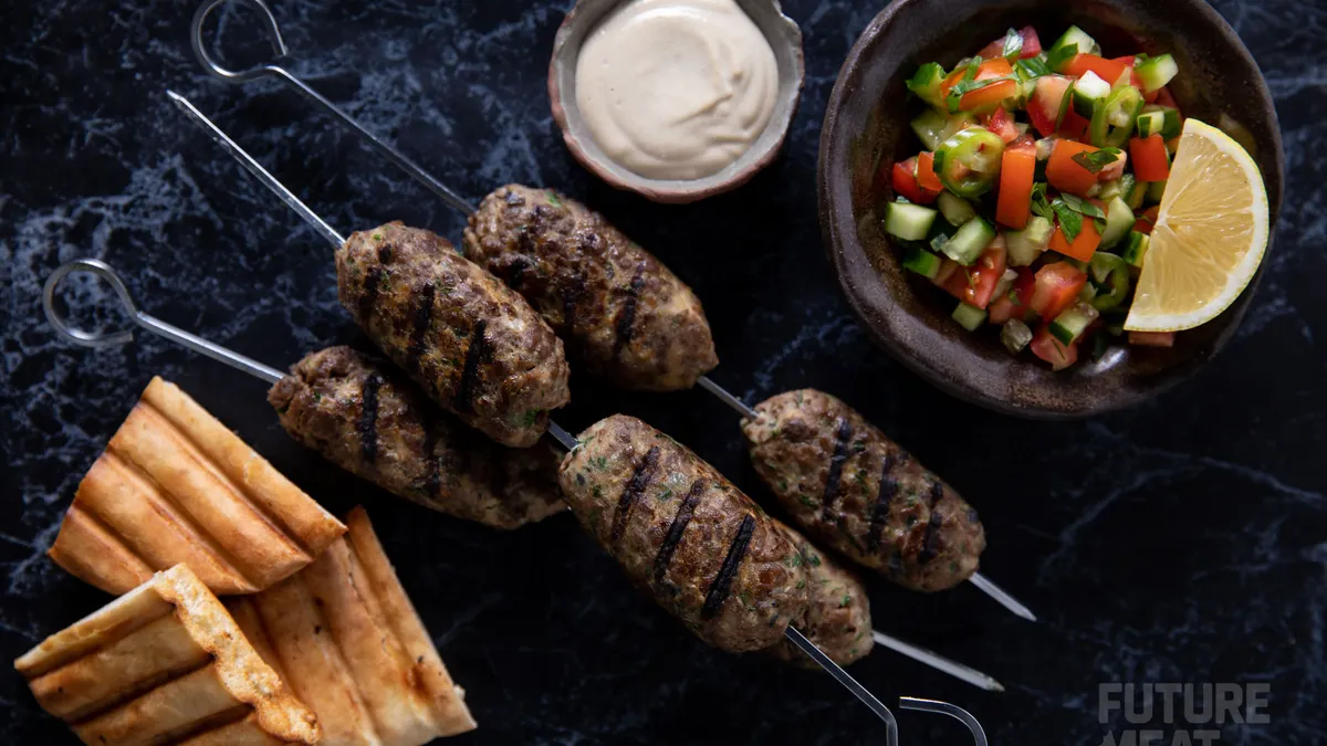 Skewers of grilled cultivated lamb pieces on a marble surface, with grilled pita triangles on one side and yogurt sauce and a tomato-cucumber-pepper vegetable garnish on the other.