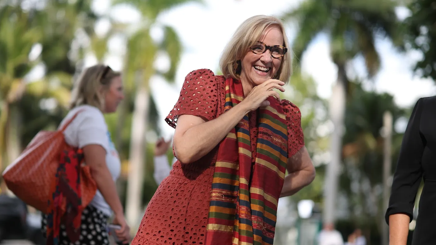 A person with blonde hair and glasses wearing a red dress surrounded by palm trees.