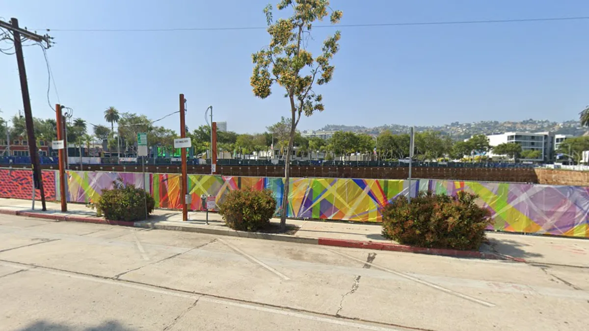 A city street with a colorful mural-lined fence blocking off a large property.