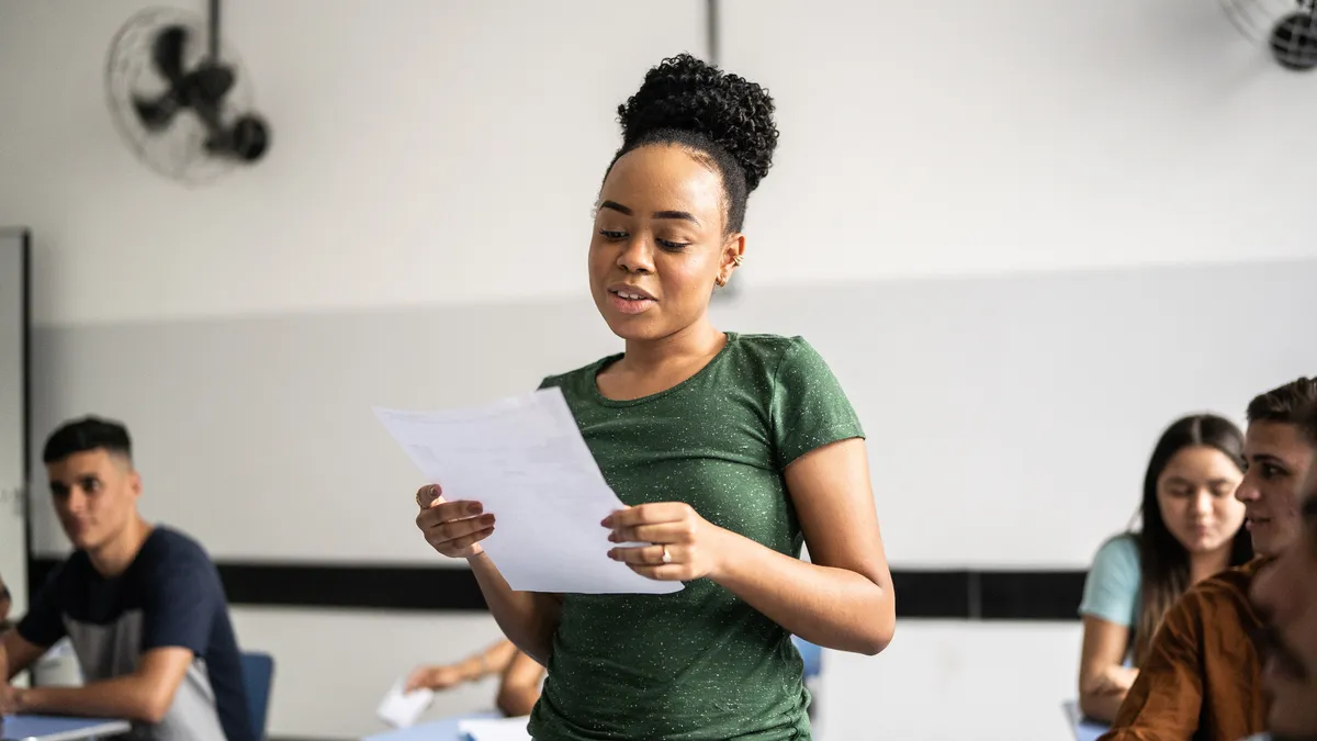 A Black teenage girl in a green shirt stands among the desks of her peers in a classroom and reads aloud from a sheet of paper.