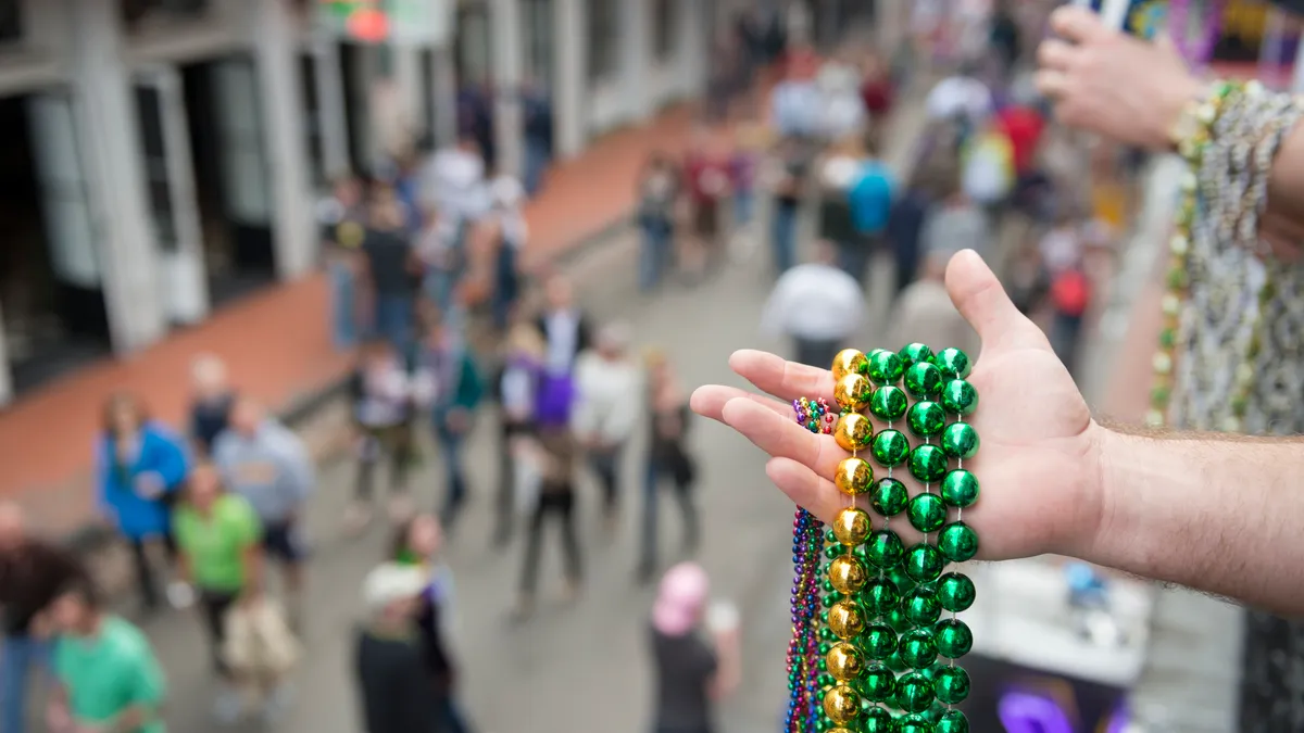 A hand holds several strands of Mardi Gras beads while standing on a balcony above Bourbon Street