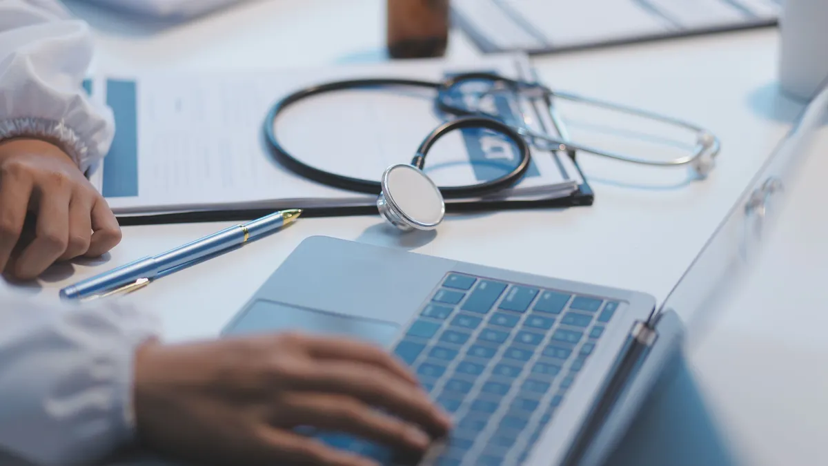 A medical professional rests their hand on a laptop keyboard while a stethoscope sits on the table next to them.