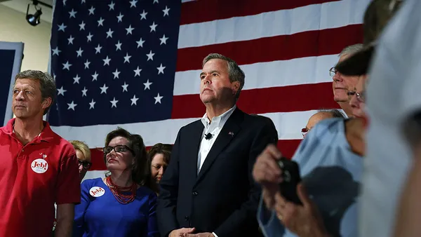 Jeb Bush is standing the center of a handful of people who have Jeb! stickers on their clothes. A large American flag is on the wall behind the people.