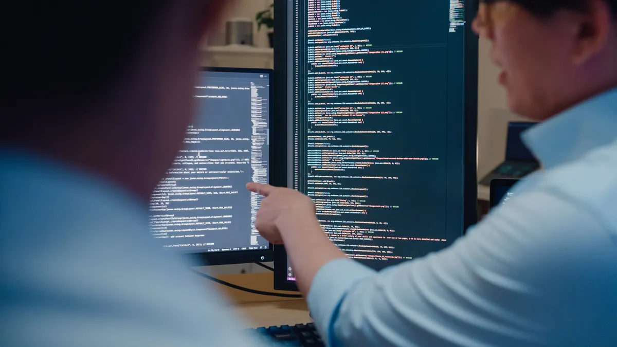 Two software developers sitting at a desk reviewing code displayed on multiple computer monitors.