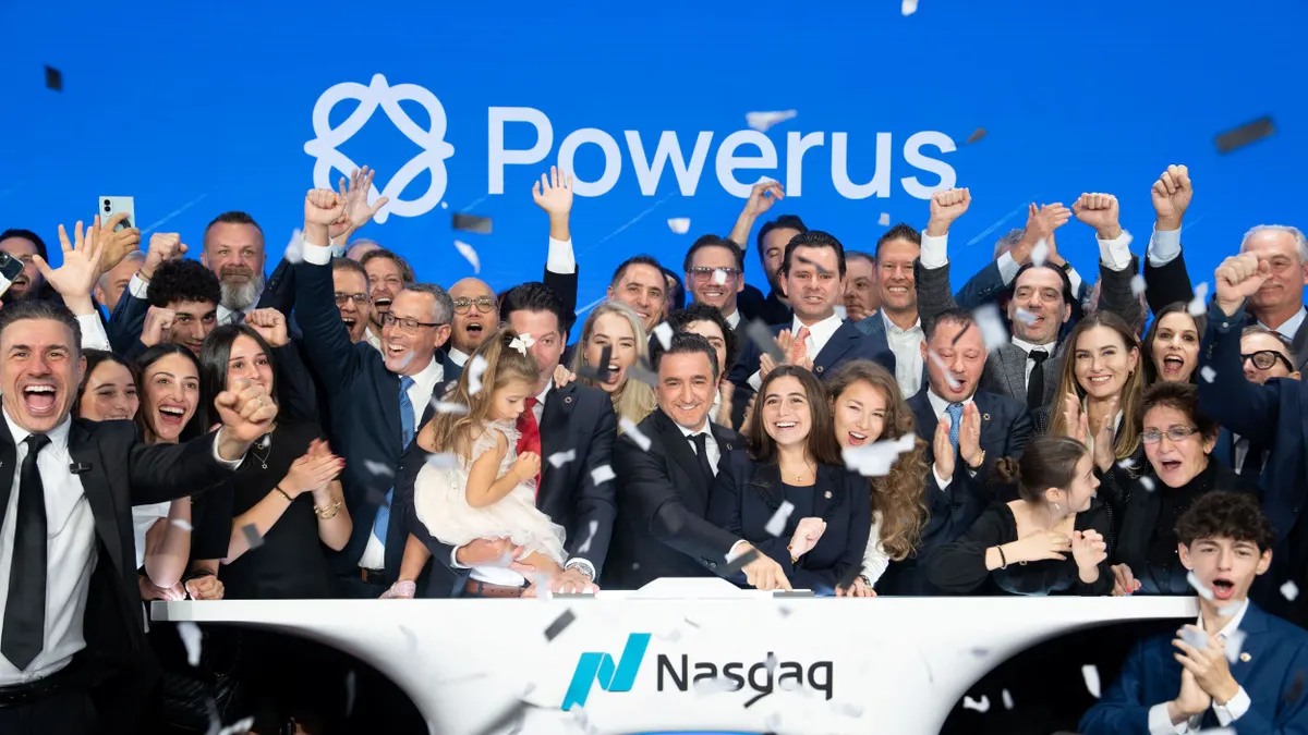 A large group of people standing behind a podium as confetti falls on them, with the blue Nasdaq screen in the background.