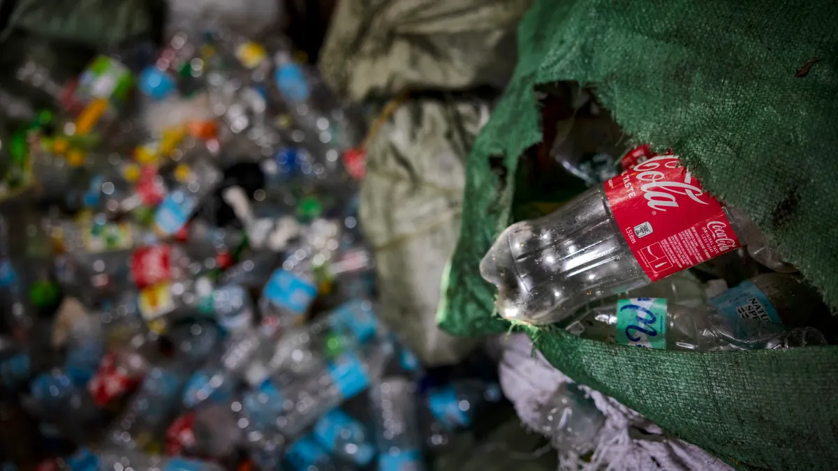 The bottom of an empty clear plastic Coca-Cola bottle sticks out from a green bag, with dozens of other empty beverage bottles for recycling blurred in the background.