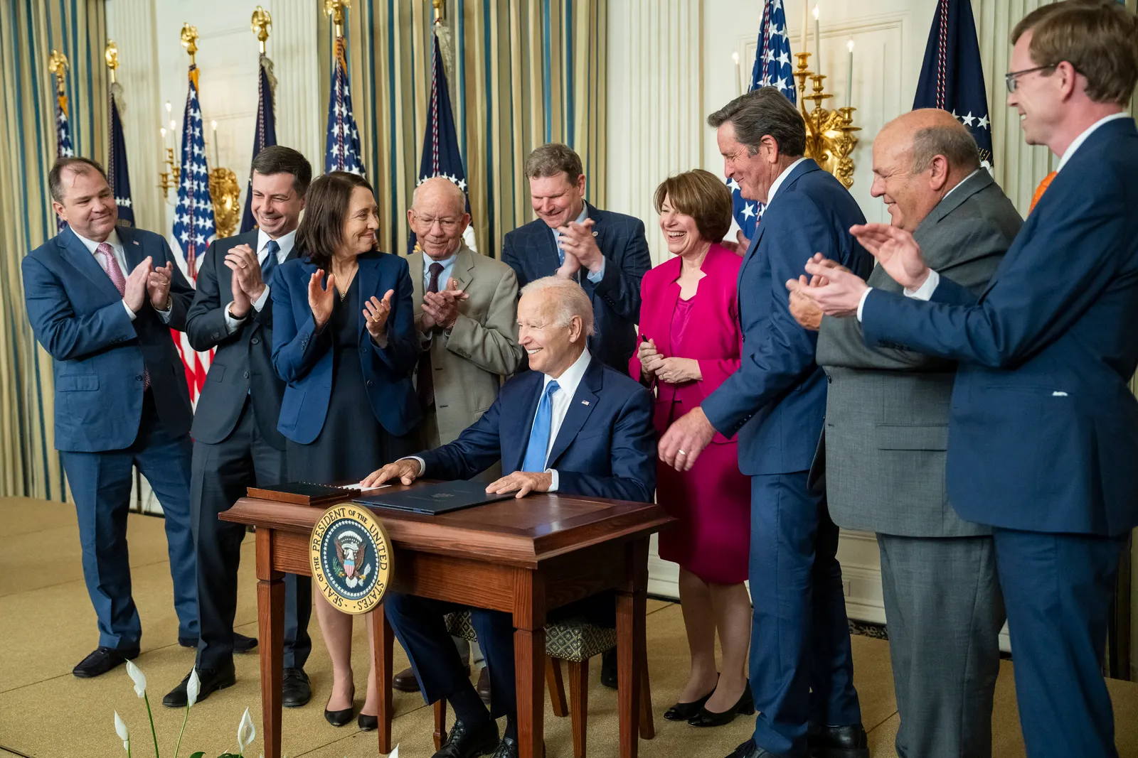 U.S. President Joe Biden sits at a desk smiling as he signs the Ocean Shipping Reform Act, surrounded by VP Kamala Harris and team