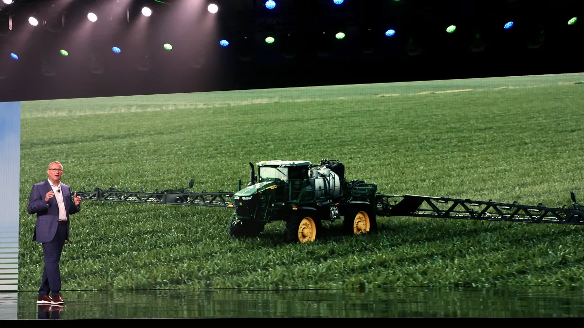 Deere's CEO is seen delivering a speech with a large picture of a tractor behind him.