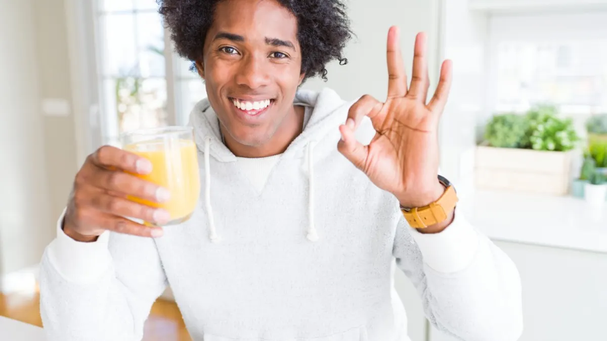 African American man holding and drinking glass of orange juice doing ok sign with fingers, excellent symbol