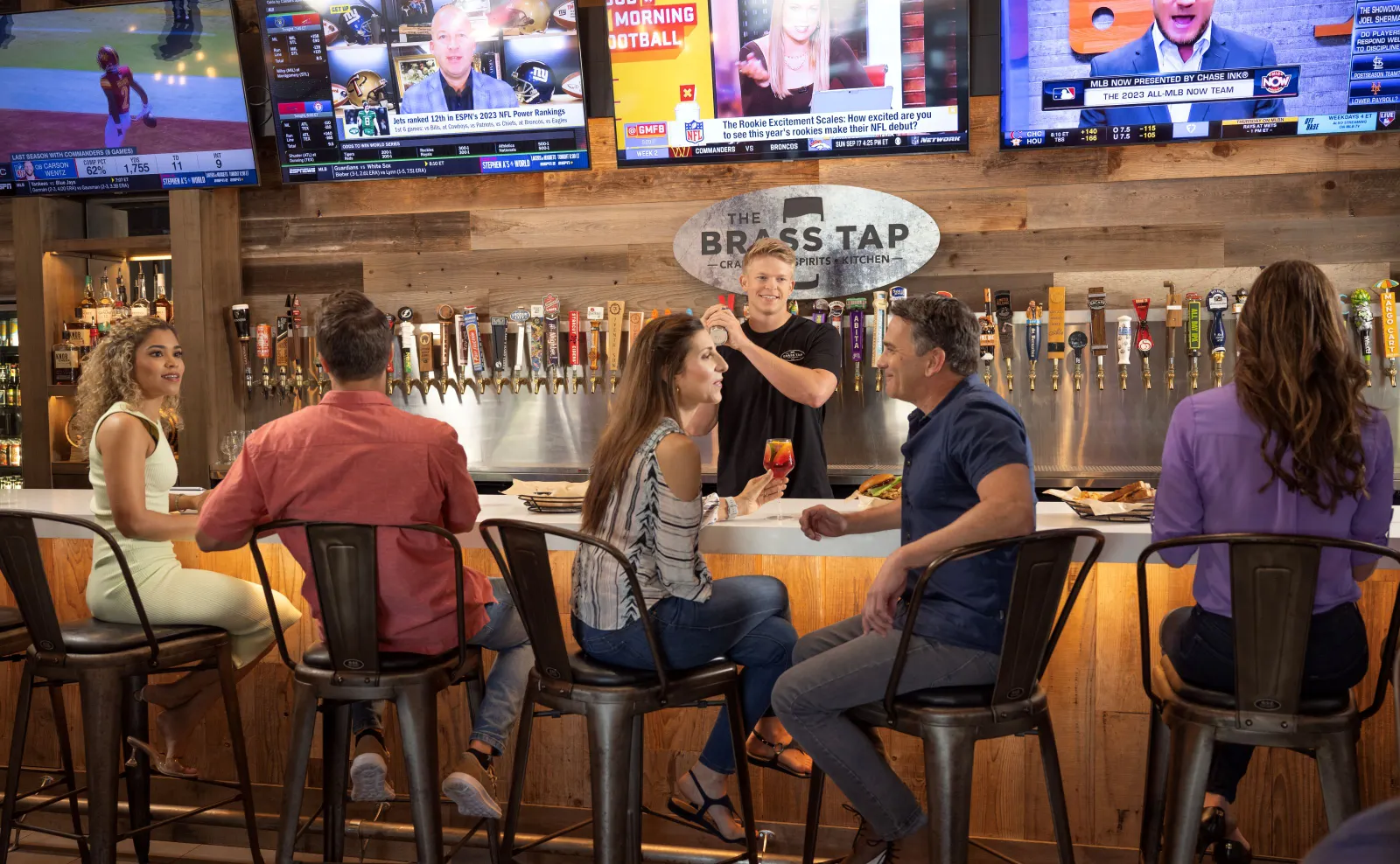 A person stands behind a bar shaking a drink and there are several people sitting at a bar talking to each other.