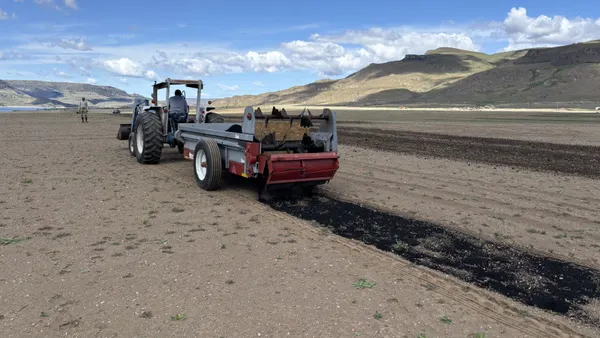 A tractor spreads dark material over a bare dirt field. A person stands supervising nearby, with mountains visible in the background.