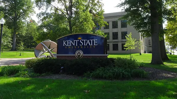 A blue sign that says "Kent State University" sits on a campus amid trees, with a three-story beige building in the middle distance.