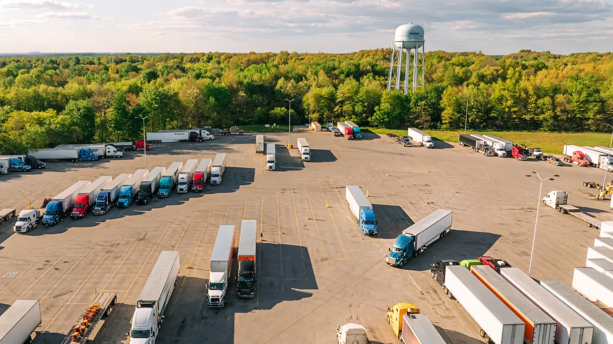 Dozens of tractor-trailers parked at a rest stop with a water tower and green trees in the background.