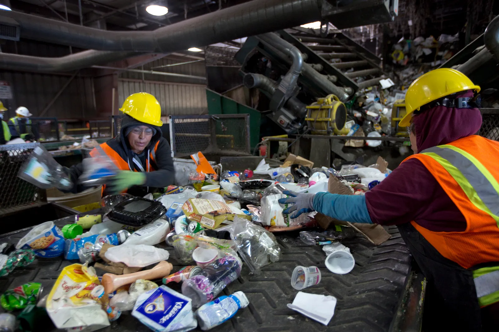 Two workers in yellow hard hats stand over a conveyor belt full of recyclable materials, primarily plastics, in an industrial facility