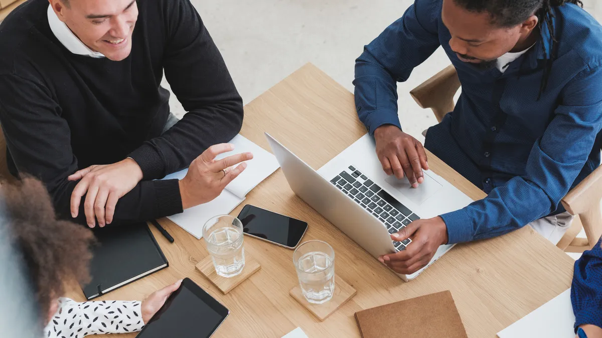 Four employees meeting at a wooden table while writing on a laptop, tablet, and notebook.