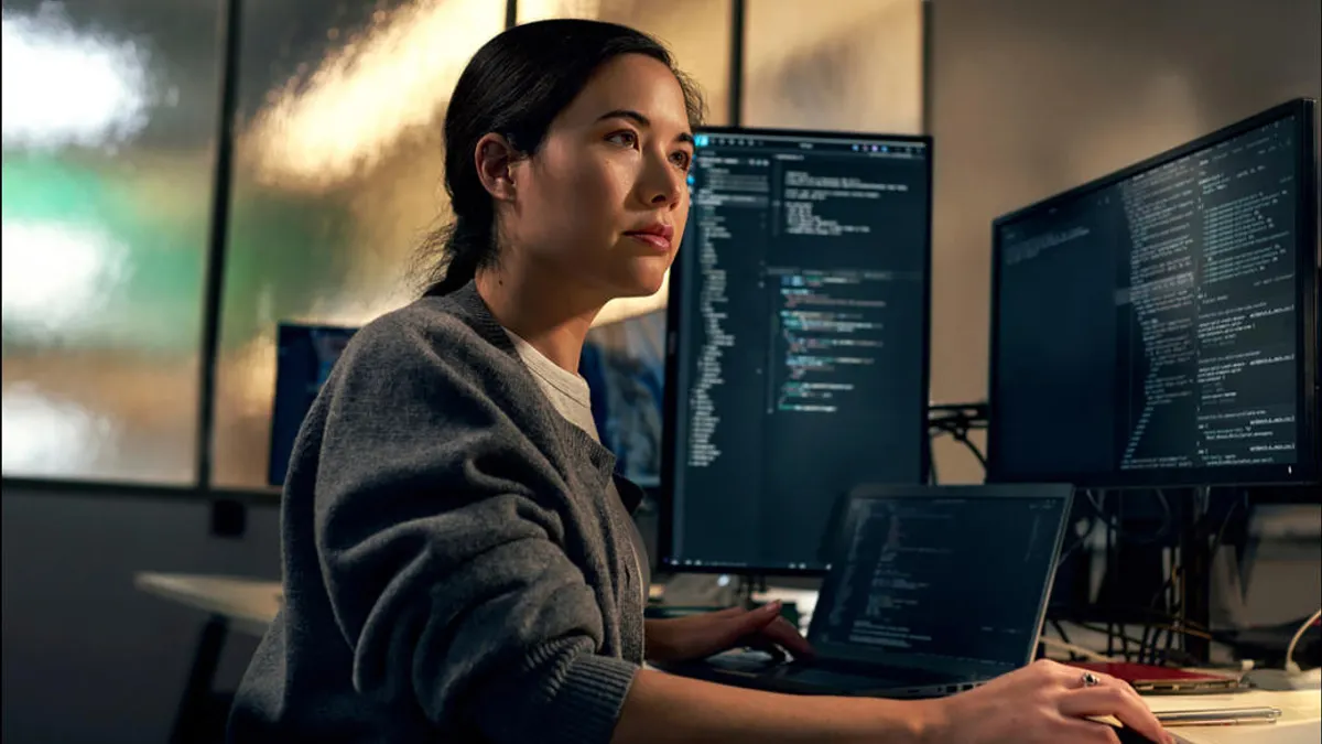 A woman in a gray sweater works at a desk with a laptop and two monitors displaying lines of computer code.