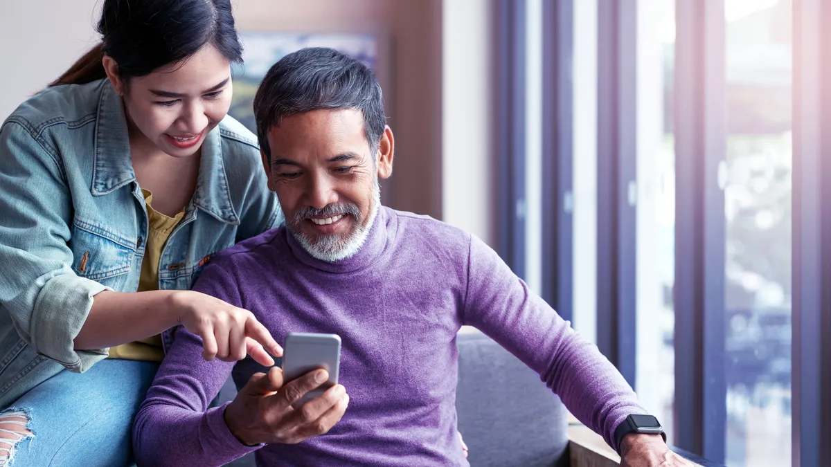 Smiling man sitting and looking at their smart phone and a woman stands behind him smiling, also looking at the phone