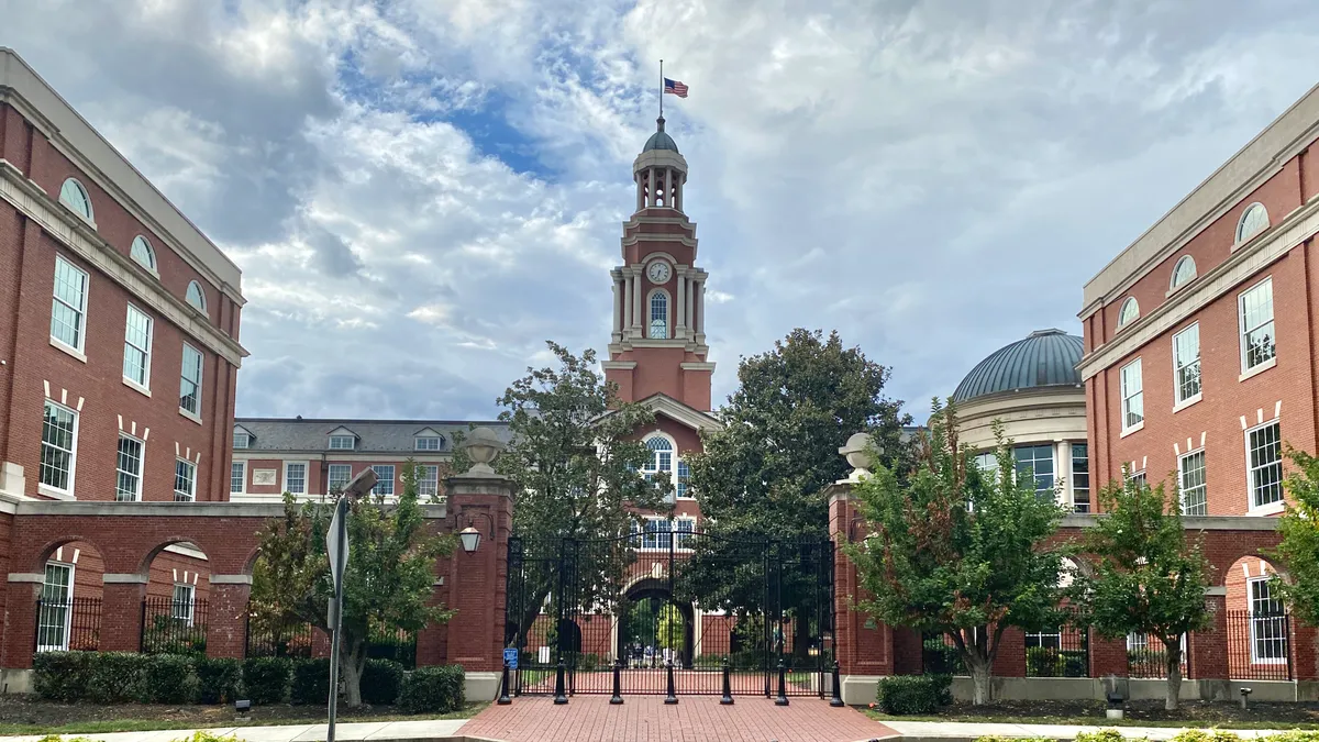 A brick courthouse with a tower and an American flag hanging from it stands on a partly cloudy day, with green trees and bushes in the foreground.