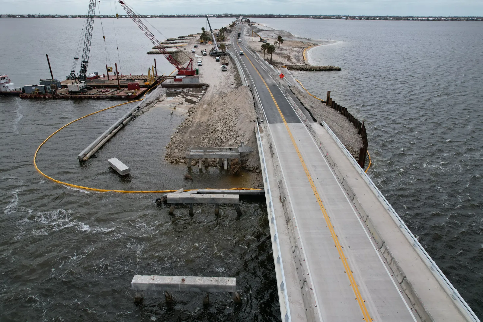 A crane and other construction equipment stands around a small island surrounded by grey water with a long road stretching across it.