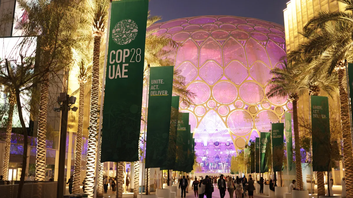 Green COP28 flags line palm trees on a walkway leading to a dome with conference attendees scattered around.