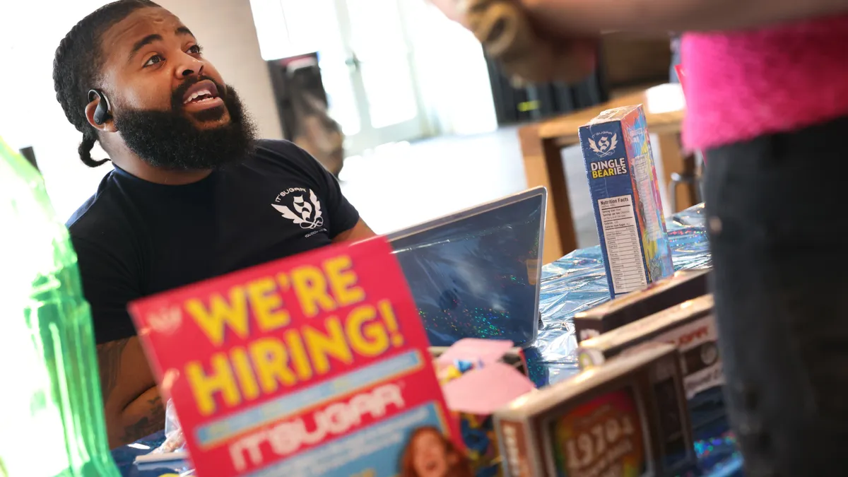 A business representative speaks with a job applicant, with a "we're hiring" sign in the foreground.