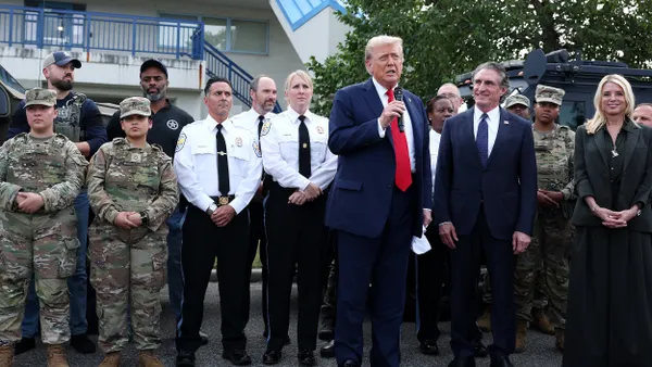 President Trump speaks into a microphone in front of people wearing military and police uniforms, a man in a suit and a woman dressed in black.
