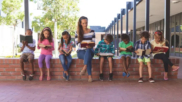 Front view of a teacher and a handful of students sitting on a brick wall outside a school building. They are all holding books.