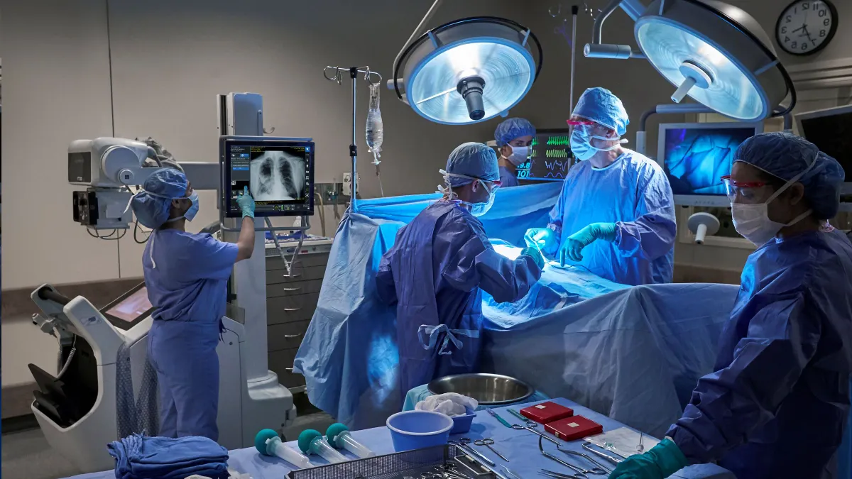 A group of people wearing blue scrubs and masks stands in an operating room. One person points to an image of lungs on a screen.
