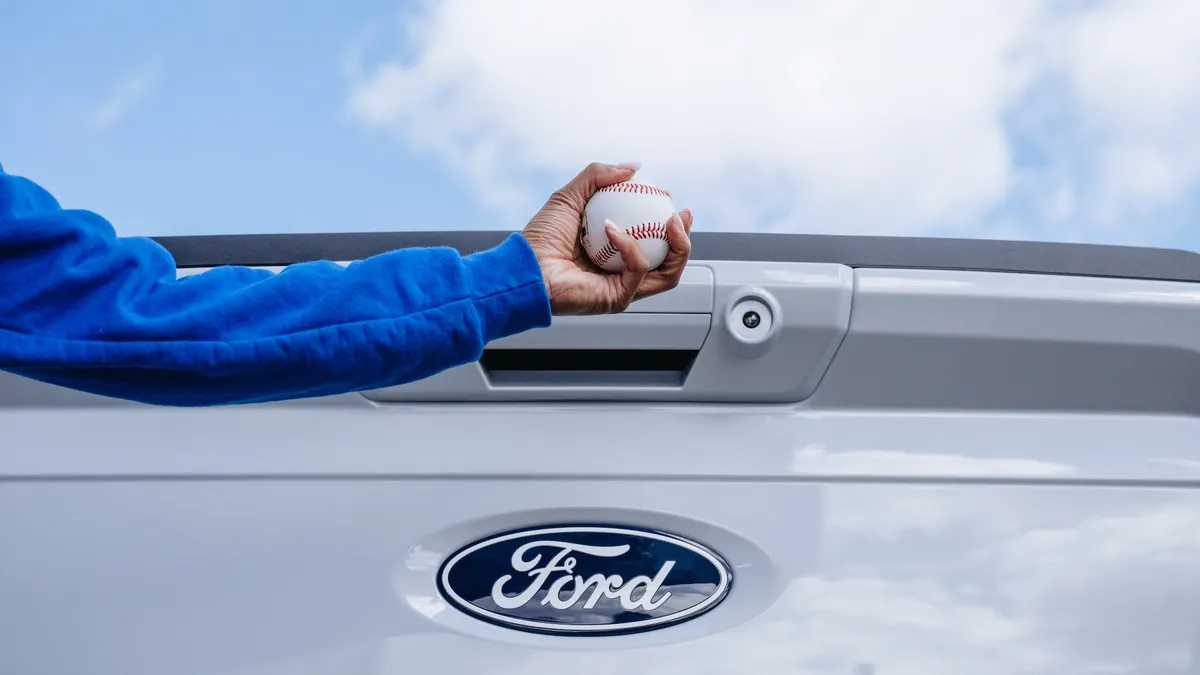 A person holds a baseball in front of a Ford truck