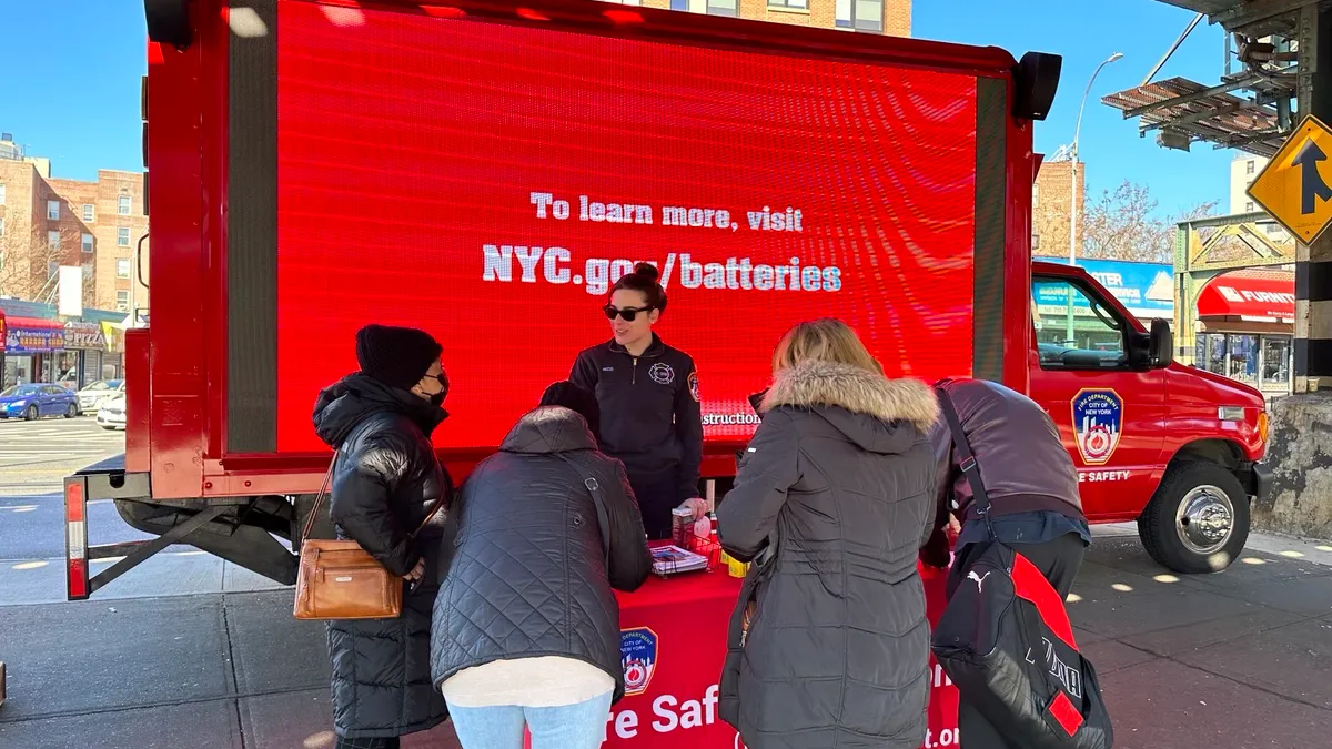 A pedestrian, EMT and fire department representative stand around a n FDNY table and van promoting lithium ion battery safety along a New York street.