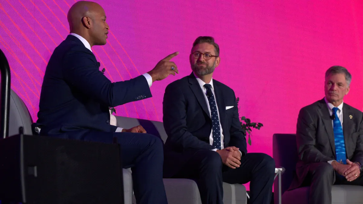 Three adults sit on chairs on a stage. They are talking. A purple screen is behind them.