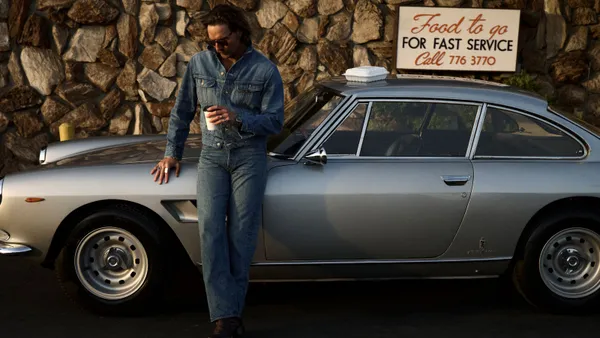 A man in a denim jacket and jeans leans against a vintage silver car and hols a cup of coffee. A styrofoam container sits on top of the car and on a wall in the background is a take-out food sign.