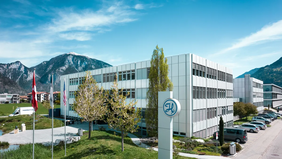 An office building is in front of mountains with a sign featuring a cursive "H."