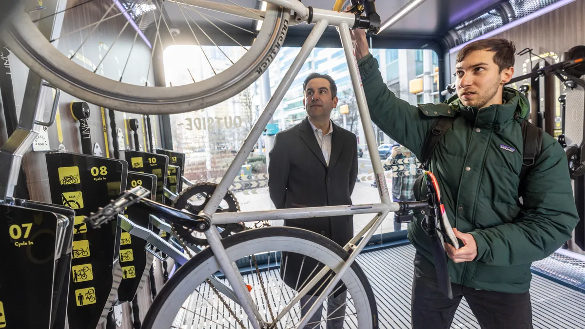 A young person holds a bike on end while pushing it into a locking stand while a public official looks on.