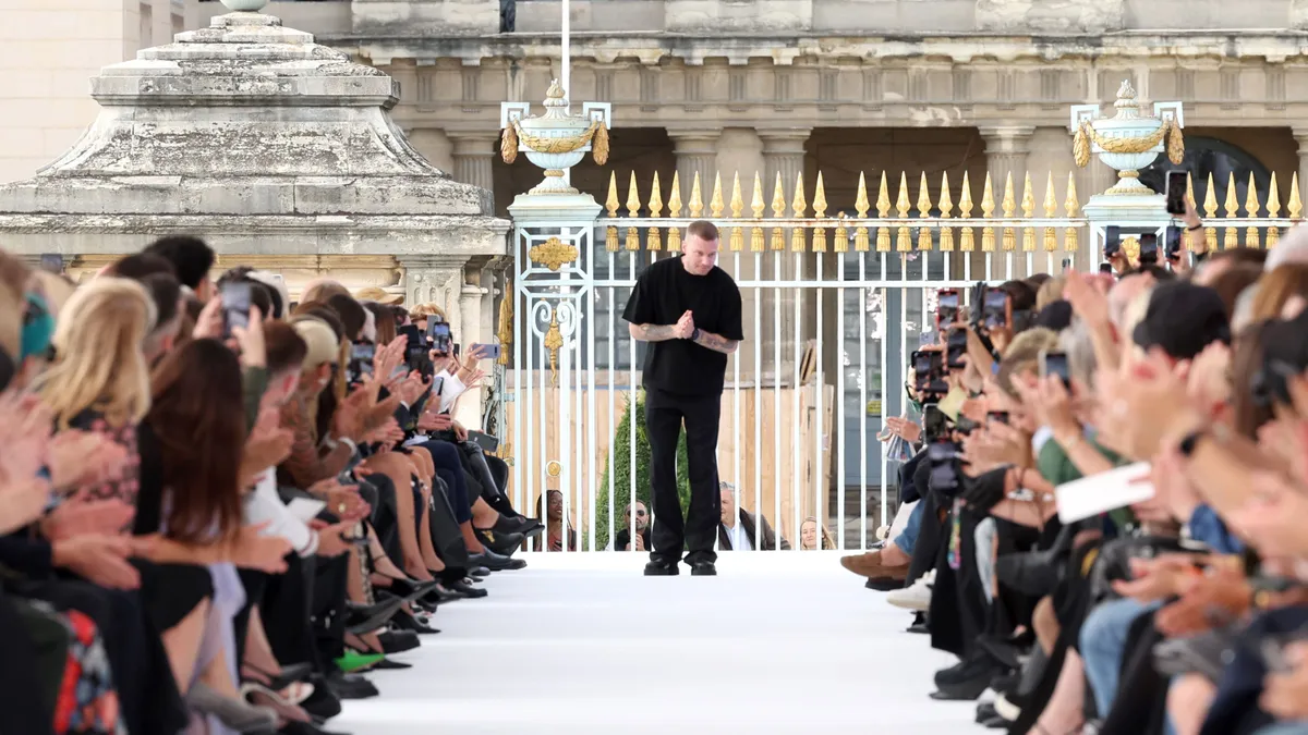 A man, designer Matthew Williams, holds his hands at the back of a fashion runway as viewers applaud from the sidelines.