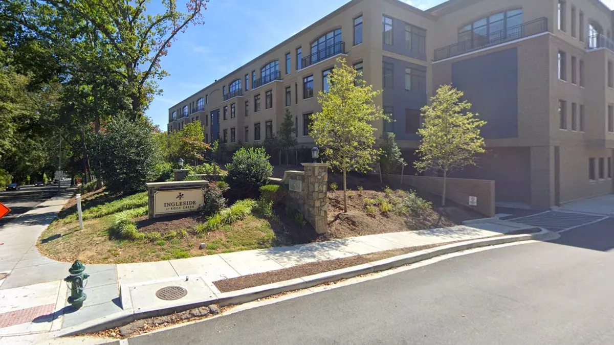 A sign for the housing complex "Ingleside at Rock Creek" is situated at a street corner, surrounded by various shrubs. Behind it stands a tan and grey multi-story building.