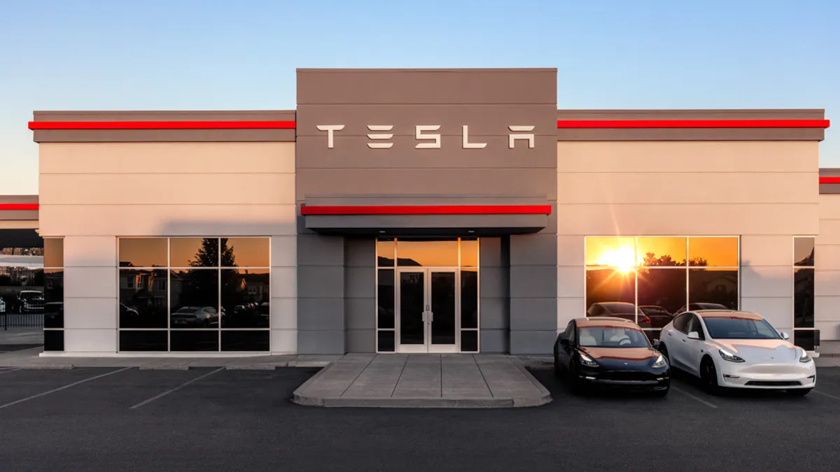 The exterior of a Tesla dealership with the sun set beaming off the window, and a blue car and a white car parked at the front.