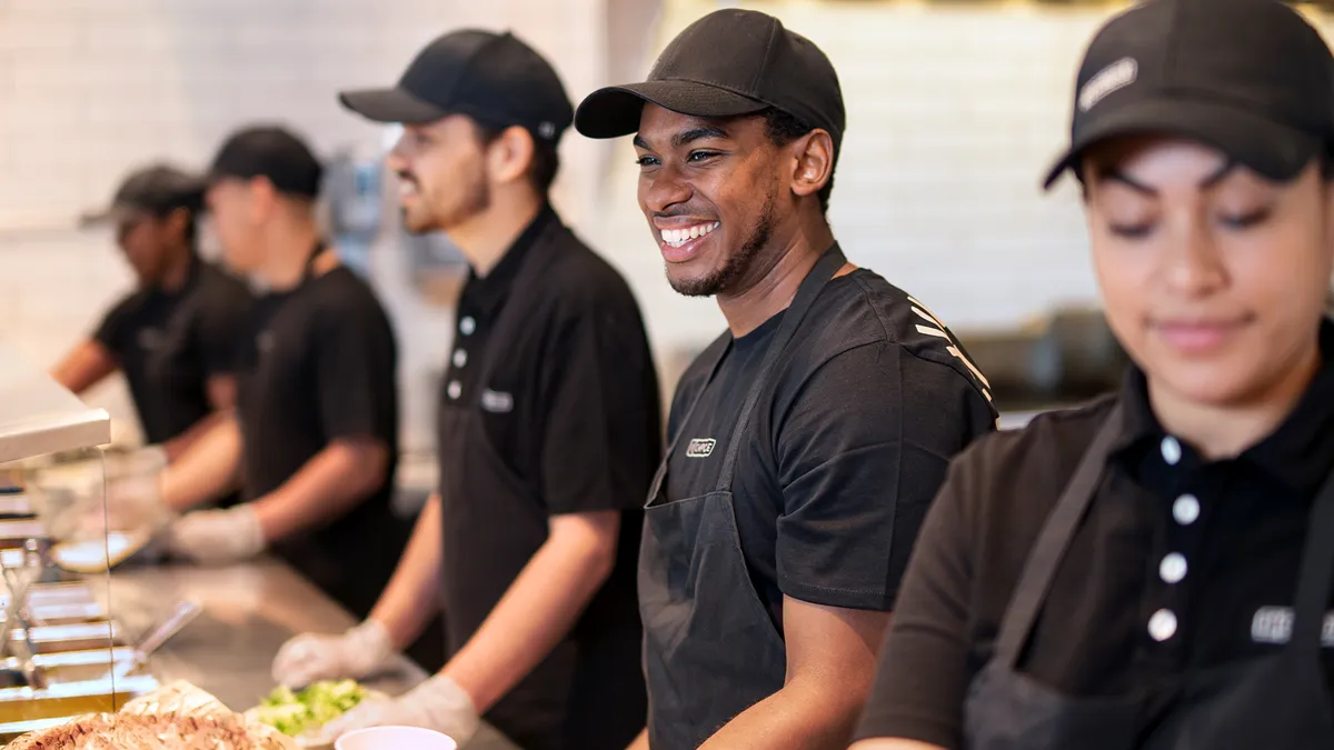 Five people in black uniforms working a makeline at a Chipotle restaurant