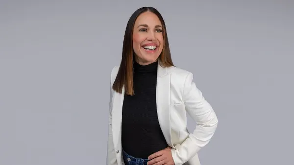 A person with long hair wearing a black shirt and white jacket is smiling.